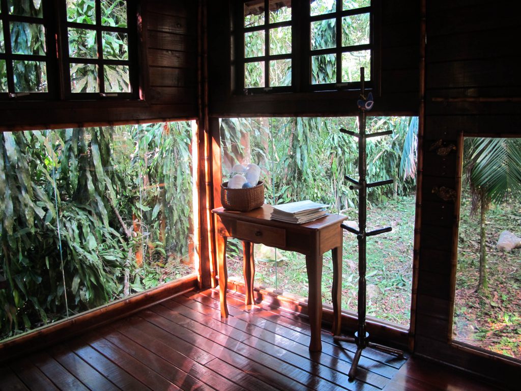 Canto de um dos bangalôs do local, com piso de madeira e mesa com toalhas. Parede de vidro com vista para a natureza.