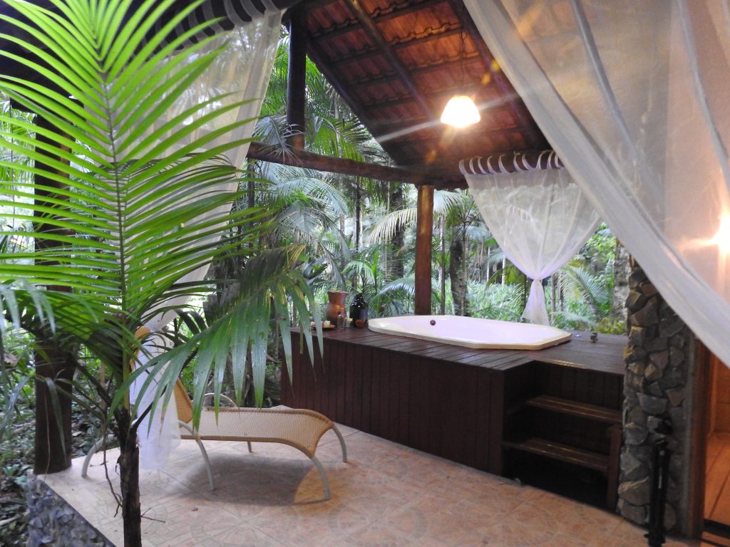 Vista do spa do glamping, com estrutura de madeira, banheira de hidromassagem, espreguiçadeira e natureza ao redor.