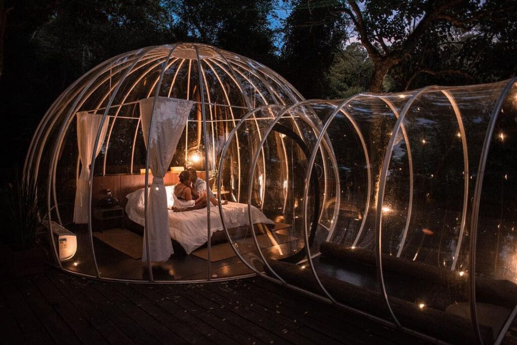 Imagem externa da bolha transparente, em uma noite, com pontos de luzes espalhados. Na cama de casal, um casal está sentado.