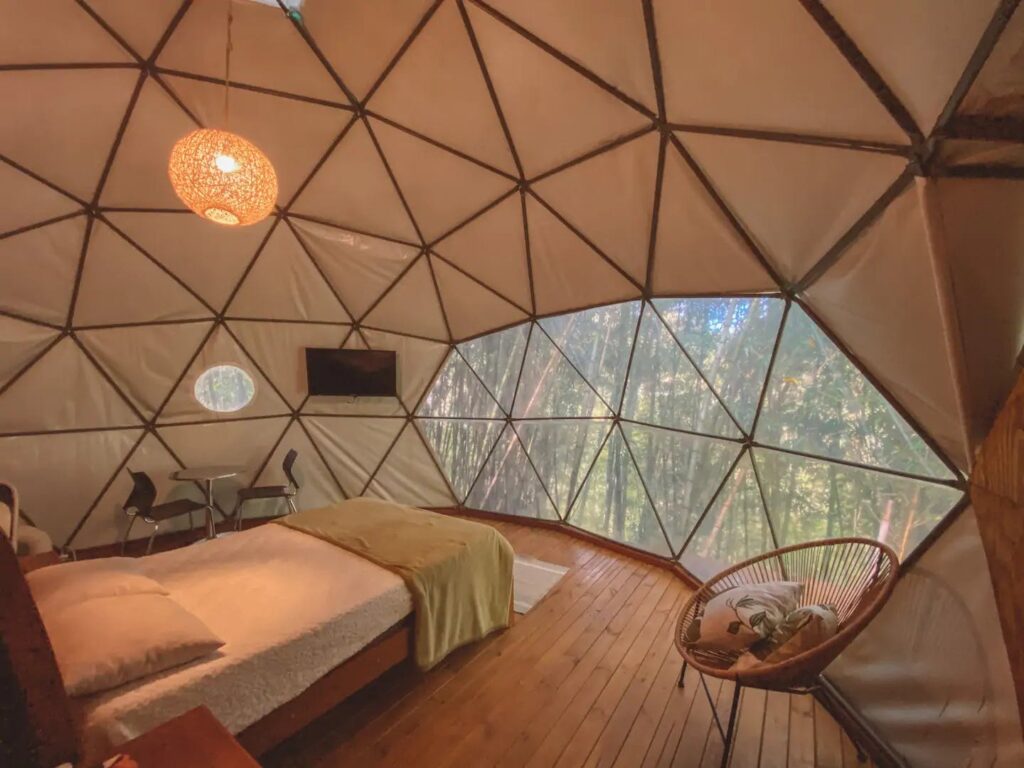 Vista interna da acomodação em formato de domo, com cama de casal, cadeira, televisão e um painel transparente para ver a natureza.