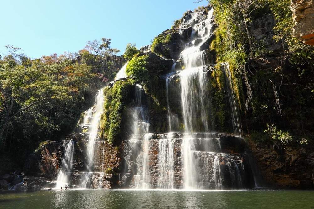 Imagem de uma cachoeira com vista de baixo, enxergando tosa sua altura, envolta por vegetação, e embaixo o poço de água.