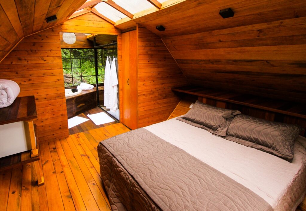 Visão interna de uma cabana na floresta do Viva Brotas Ecoparque, com cama de casal e banheira com vista para a mata.